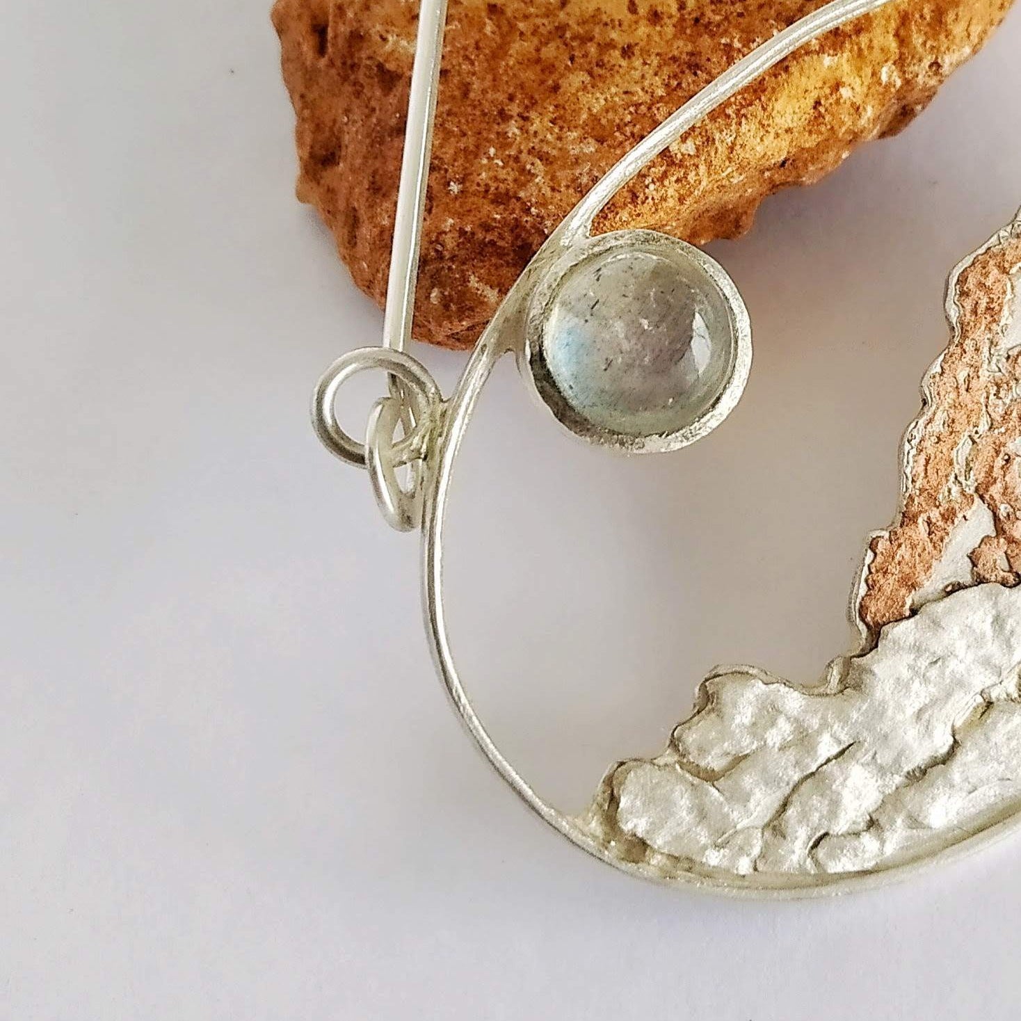 A detail of a silver pendant with copper and a moonstone representing a landscape under the moon