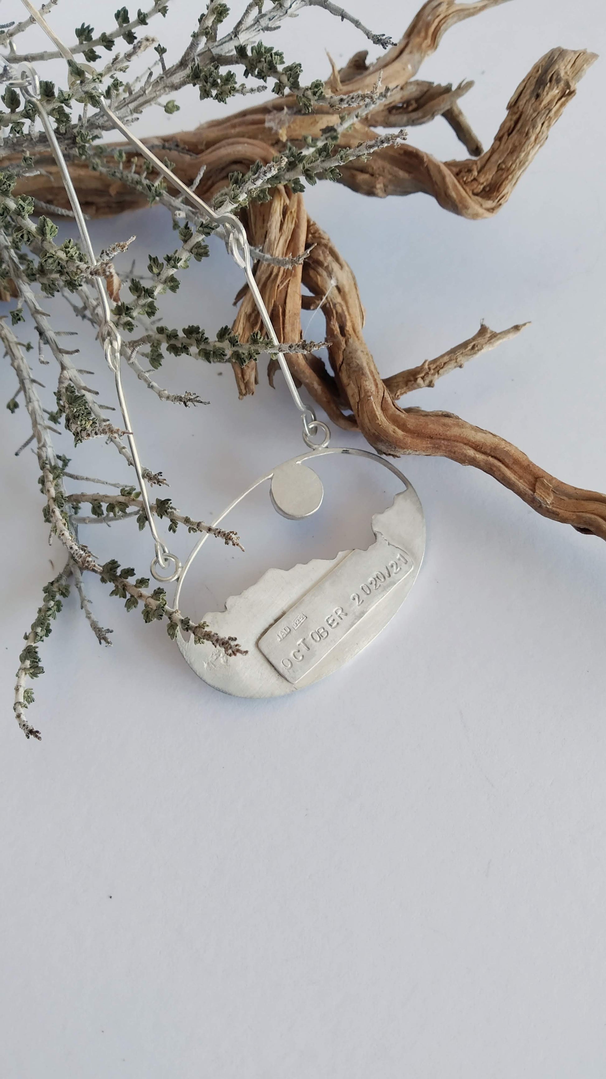 The back of a silver pendant with a labradorite (moonstone) and a copper representing a landscape under the moon. On a silver tag is imprinted October 2020/21