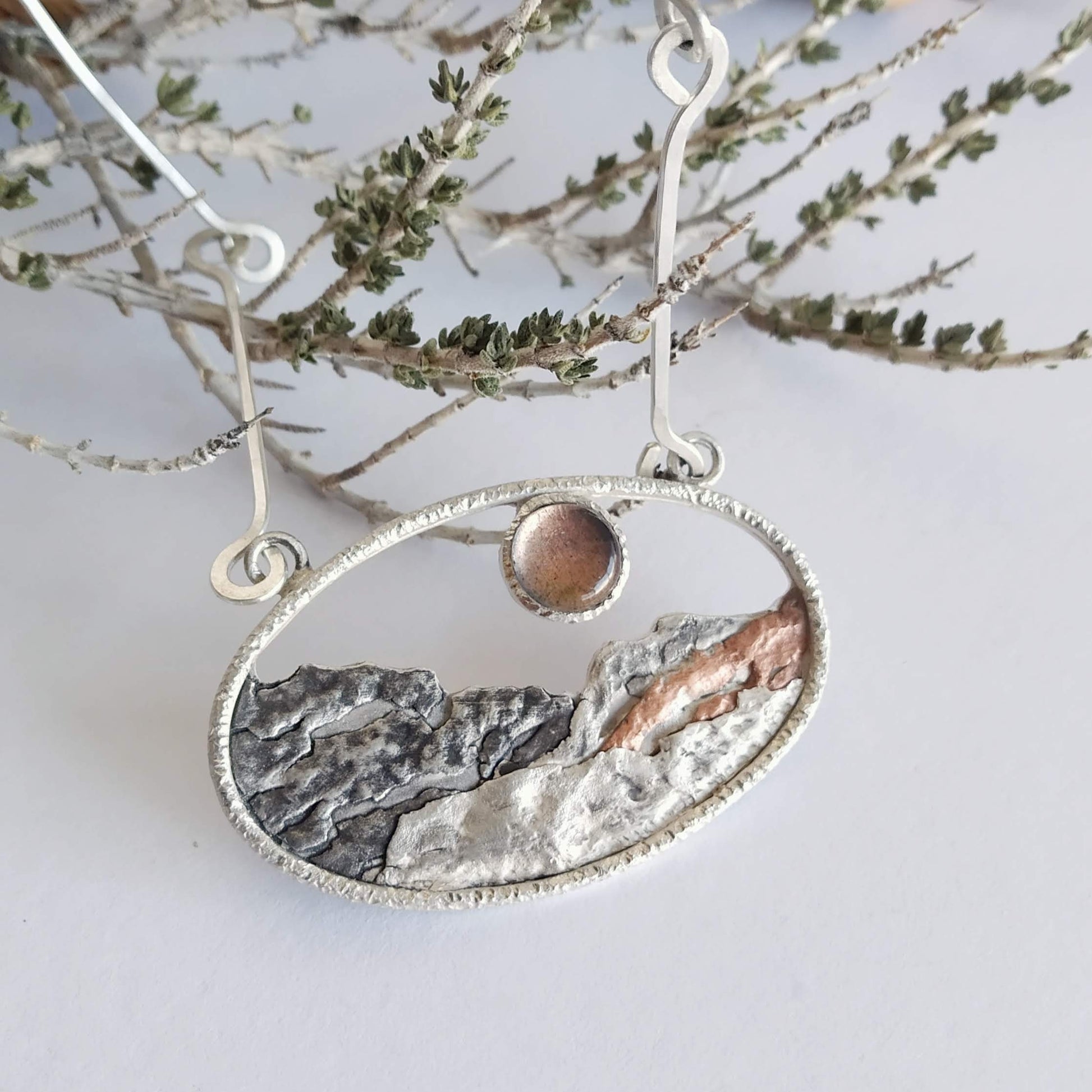 A silver pendant with a labradorite and some copper, representing a landscape at sunset