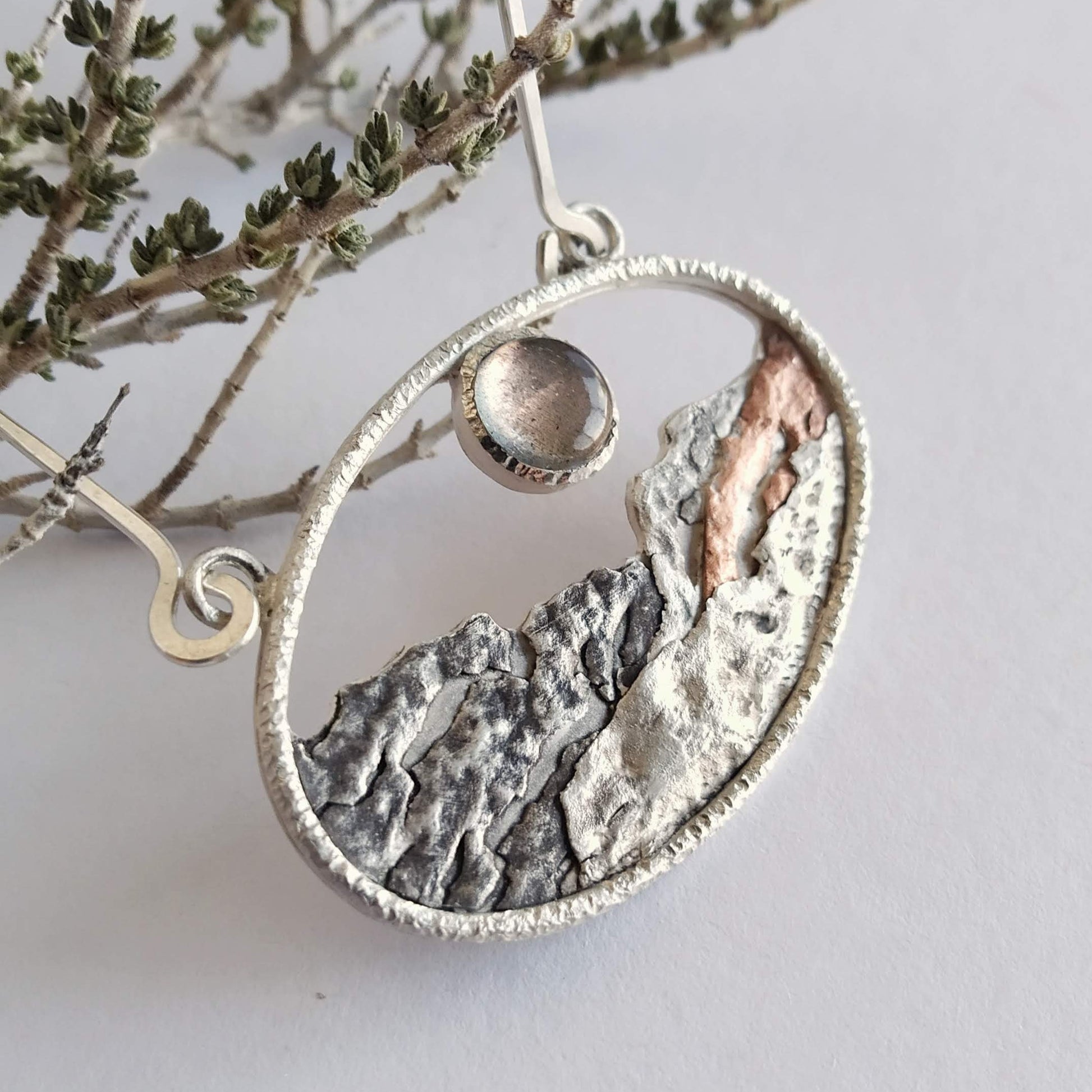 A silver pendant with a labradorite and some copper, representing a landscape at sunset