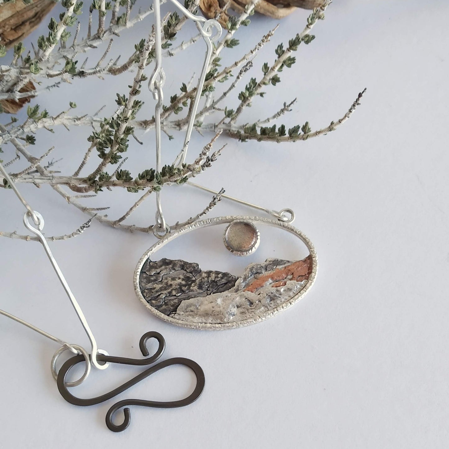 A silver pendant with a labradorite and some copper, representing a landscape at sunset. It has a silcer chain ad an S-shaped clasp